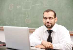 A smiling teacher with his laptop open on his desk.
